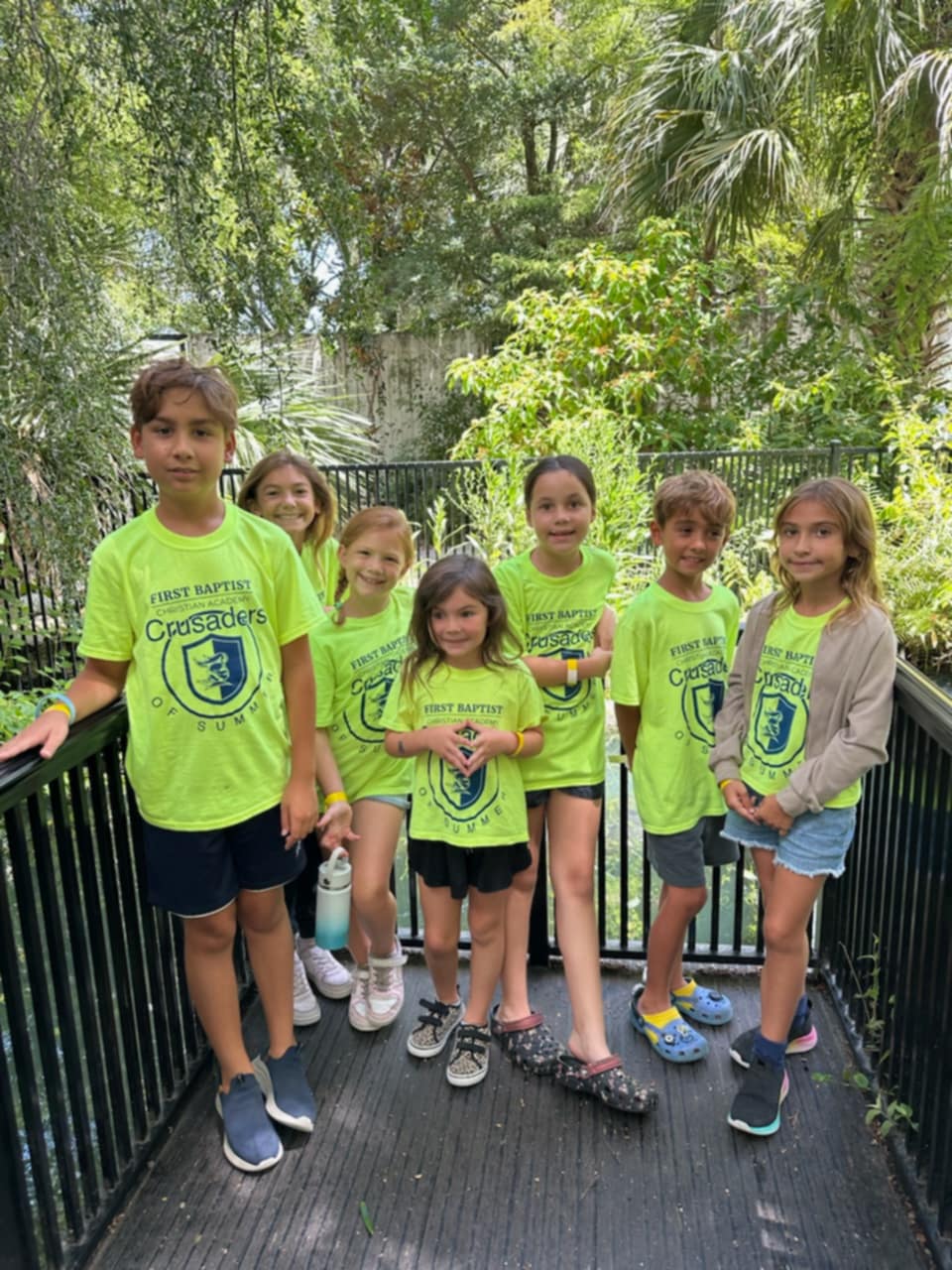 A group of elementary-aged students wearing bright green First Baptist Christian Academy summer shirts stand together on a wooden boardwalk, smiling outdoors with lush greenery and trees surrounding them.